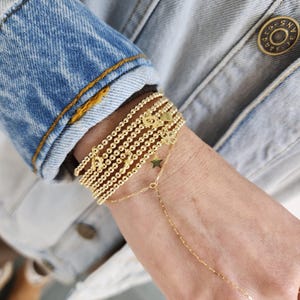 May include: A stack of gold beaded bracelets with charms, including a star and a heart, on a person's wrist. The bracelets are layered on top of each other and are secured with a clasp.