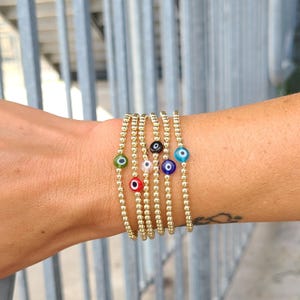 Könnte beinhalten: Goldene Perlenarmbänder mit bunten Evil-Eye-Anhängern. Die Armbänder bestehen aus kleinen, runden Goldperlen und haben Anhänger in Grün, Rot, Schwarz, Rosa, Blau und Weiß. Die Armbänder sind an einem Handgelenk gestapelt.