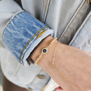 May include: Gold beaded bracelet with a blue evil eye charm. The charm is set with small clear stones.