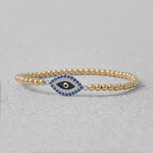 May include: A gold beaded bracelet featuring a blue evil eye charm. The charm is set with small white stones.