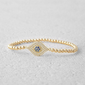 May include: A gold beaded bracelet featuring a blue and white evil eye charm. The charm is made of cubic zirconia and has a blue centre.