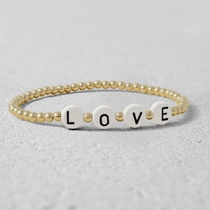 May include: A gold beaded bracelet with white beads that spell out the word "LOVE".