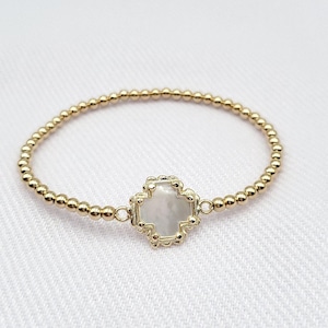 May include: A gold beaded bracelet with a white, cross-shaped charm. The charm has a decorative, ornate design.