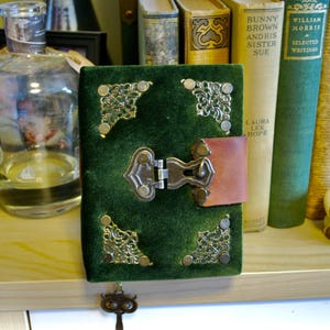 May include: A green velvet journal with ornate metal corner accents and a clasp. The journal has a brown leather closure and a decorative key charm. Books and a glass bottle are in the background.