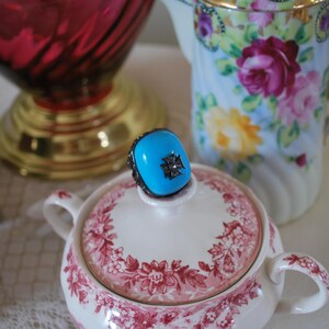 May include: A turquoise ring with a silver filigree setting and a black cross design sits atop a floral-patterned sugar bowl. A floral-decorated teapot and a red glass vase are in the background, suggesting a vintage or antique theme.