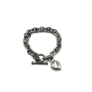 May include: A silver-toned chain link bracelet featuring a toggle clasp closure and a single heart-shaped locket charm. The bracelet is constructed from a series of interconnected circular metal links with a polished finish and a dark patina.