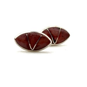May include: A pair of oval-shaped cufflinks with a silver-colored border. The cufflinks have a reddish-brown center with a silver-colored V-shaped design. The background is white.