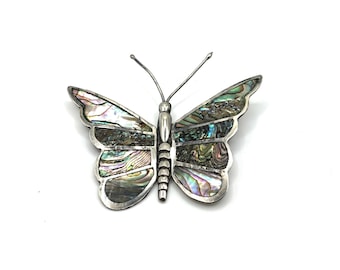 May include: A silver butterfly brooch with iridescent abalone shell wings. The brooch has a detailed design with a silver body and antennae. The wings display a range of colors, including green, blue, and purple.
