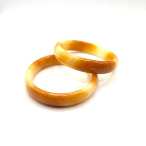 May include: Two smooth, circular bangle bracelets crafted from a translucent, polished stone material. The bangles feature a gradient of warm, earthy tones, transitioning from a deep, saturated amber-orange to a lighter, creamy yellow hue.