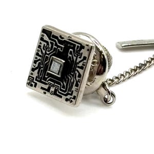 May include: Silver-toned tie tack with a square, circuit board design. The square has a black circuit pattern with a central square and is attached to a chain and a pin. The tie tack is on a white background.