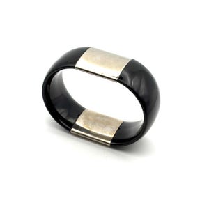 May include: A black bangle bracelet with two silver-colored rectangular accents. The bracelet is a solid, rounded shape, and the silver accents are placed on opposite sides. The bracelet is set against a white background.