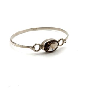 May include: A silver bangle bracelet featuring an oval, smoky quartz gemstone. The gemstone is set in a silver bezel and connected to the bracelet with small silver loops. The bracelet has a simple, elegant design.