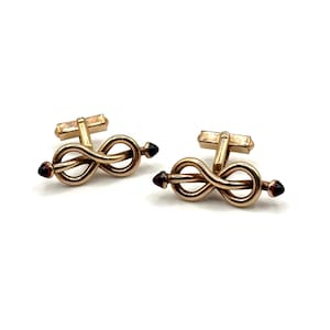 May include: A pair of gold-toned cufflinks featuring an infinity knot design. Each cufflink has a rectangular bar and is accented with a dark red, pointed stone at each end. The cufflinks are set against a white background.