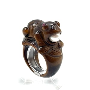 May include: A brown tiger's eye ring with a carved frog design. The frog has a pearl in its mouth and a red stone for an eye.
