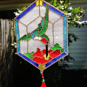 May include: A colorful beaded hummingbird hanging ornament with a hexagonal shape and a red bell at the bottom. The ornament is made of small beads in shades of red, green, blue, and white. The text "K Wynn Jewelry Hexagon Hummingbird Pattern" is visible at the bottom of the ornament.