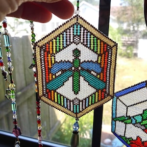 May include: A colourful beaded dragonfly hexagon suncatcher with a rainbow border and a white background. The suncatcher is hanging from a window and is made of beads. The text "K Wynn Jewelry Dragonfly Hexagon Sun Catcher" is at the bottom of the image.