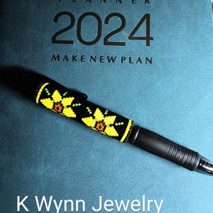 May include: A black pen with a beaded design featuring yellow and brown flowers. The pen is lying on a teal planner with the text "PLANNER 2024 MAKE NEW PLAN" and "K Wynn Jewelry".