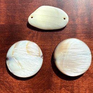 May include: Three iridescent, off-white shell pendants and beads. One is an oblong shape with two holes, and the other two are round. The shells have a pearlescent sheen and are set against a dark brown wooden surface.