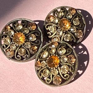 May include: Three ornate, round buttons with a bronze-colored filigree design. Each button features a large, amber-colored center stone surrounded by smaller, clear and amber-colored crystals. The buttons are on a pink surface.