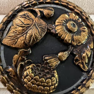 May include: A round, antique button with a black center and bronze-colored floral and leaf design. The button features a detailed relief of a flower, leaves, and a pinecone-like element, surrounded by a textured bronze border. The button is likely made of metal.