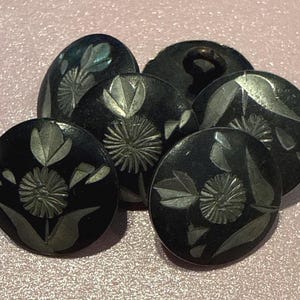 May include: A collection of six black buttons with engraved floral designs. The buttons vary in shape, including oval and round, and feature detailed silver-colored flower and leaf patterns. The buttons are arranged on a textured pink surface.