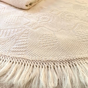 May include: An off-white textile featuring a raised, textured geometric pattern across its surface. The fabric is folded at the top and finished with a long, knotted fringe along the bottom edge, creating a decorative border detail.