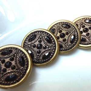 May include: Four antique brass buttons with a detailed design. Each button features a dark brown center with a pattern of raised dots and ovals, surrounded by a gold-colored rim. The buttons are arranged in a diagonal line.