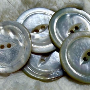 May include: A collection of four round, iridescent mother-of-pearl buttons. Each button features a smooth, polished surface with a natural pearlescent sheen and a central recessed oval area containing two small holes for attachment purposes.