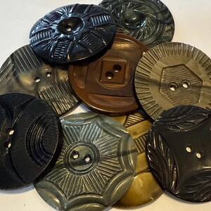 May include: A collection of vintage buttons in various shades of brown, black, and gray. The buttons have different carved designs, including geometric patterns and floral motifs. Some buttons have two or four holes for sewing.