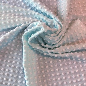 May include: A light blue fabric with a white polka dot pattern. The fabric has a textured, raised surface.