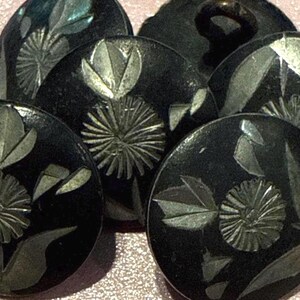May include: A collection of round black buttons featuring an inlaid silver-toned floral design. Each button displays a central flower with radiating petals and surrounding leaves, with a visible shank on the back of one button for attachment.