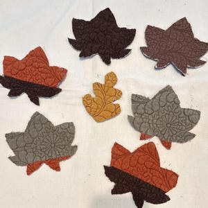 May include: A collection of fabric leaf appliqués in various colors, including brown, orange, and gray. The leaves have a textured, quilted appearance and are arranged on a white background. The leaves are in different shapes and sizes, suggesting a fall or autumn theme.