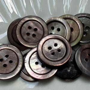 May include: A collection of round, four-hole buttons with a dark, iridescent finish. Each button features a smooth, slightly recessed center and a raised outer rim, displaying subtle shifts of deep brown, purple, and charcoal tones in the light.