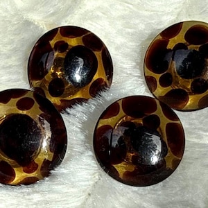 May include: Four round buttons with a dark brown and gold spotted design. The buttons have a glossy finish and are arranged on a white, textured surface. These buttons could be used for sewing or crafts.