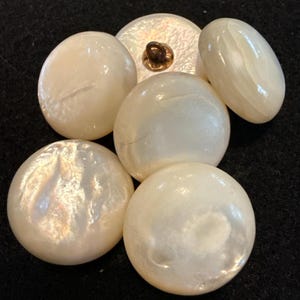 May include: A collection of iridescent, off-white buttons in various shapes and sizes. The buttons include round, oval, and a flat button with a gold-colored shank. The buttons have a pearlescent sheen, suggesting a mother-of-pearl or similar material.