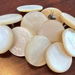 May include: A collection of iridescent, pearlescent buttons in various shapes and sizes. The buttons are primarily oval and round, with a creamy white color and a subtle sheen. Some buttons have a brass shank.