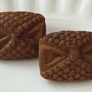 May include: Two small, square, wooden objects with intricate carved designs. The objects are a warm brown color and feature a basket weave pattern on the top and sides, with a bow-like design on the front. They are likely decorative or functional items.