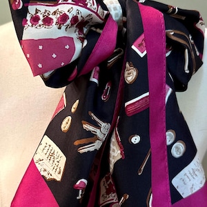 May include: A black silk scarf featuring a vibrant print of various objects including keys, coins, handwritten notes, and floral patterns, finished with a bold magenta border. The scarf is tied in a knot, displaying the intricate multi-colored graphic design.