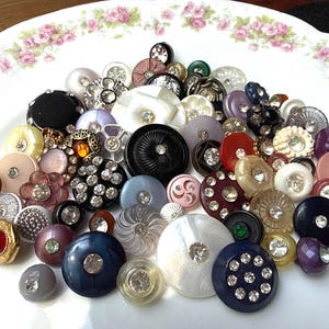 May include: A collection of vintage buttons in various shapes, sizes, and colors, including black, white, red, and blue. Many buttons feature rhinestones or other decorative elements. The buttons are arranged on a white plate with a floral border.