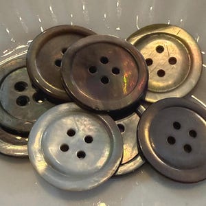 May include: A collection of round, four-hole buttons in various iridescent shades of gray, brown, and cream. Each button features a smooth, polished surface with a distinct sheen, showcasing natural color variations and a raised outer rim.