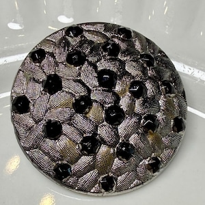 May include: A circular button featuring a textured, metallic silver woven pattern across its surface. The design is accented with multiple small, faceted, black, round stones embedded throughout the intricate, grid-like metallic structure.