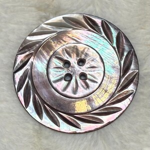 May include: A round, iridescent button with a dark, pearlescent finish. The center features a circular section with four holes and a radial pattern, surrounded by an outer ring decorated with a carved, leaf-like motif around the edge.