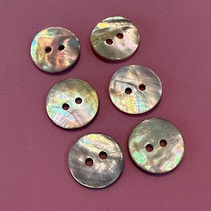 May include: Six iridescent, round buttons with two holes each are arranged on a solid, dark pink background. The buttons have a pearlescent sheen, reflecting colors like green, gold, and purple.