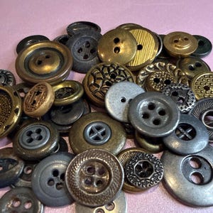 May include: A collection of assorted vintage buttons in various sizes and designs. The buttons are primarily in shades of gold, bronze, and silver, with some featuring intricate patterns and textures. The buttons have two or four holes.