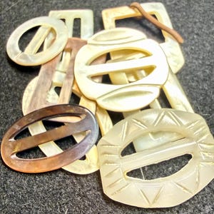 May include: A collection of assorted belt buckles in various shapes and sizes, including round, oval, and rectangular designs. The buckles feature smooth, iridescent, and carved surfaces in shades of cream, beige, and dark brown.