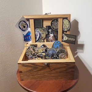 May include: A wooden box with a hinged lid, filled with a variety of military-themed challenge coins and patches. The box is decorated with a photo of a young boy in a military uniform, and several patches featuring military insignia.