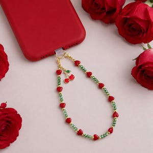 May include: A red smartphone case attached to a beaded phone charm featuring small green faceted beads, gold-tone spacer beads, and red heart-shaped beads. A gold-tone metal charm shaped like two red cherries with green leaves hangs from the clasp.