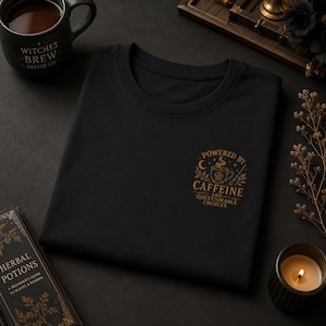 May include: A black t-shirt featuring a gold graphic on the chest. The design includes a coffee cup with a pentagram, a crescent moon, stars, and the text "POWERED BY CAFFEINE AND QUESTIONABLE CHOICES" written in a decorative, serif font style.