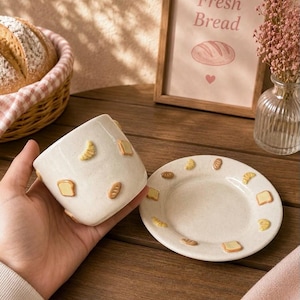 Könnte beinhalten: Eine Keramiktasse und eine passende Untertasse in cremeweißer Sprenkeloptik, beide verziert mit kleinen, erhabenen, dreidimensionalen Tonapplikationen verschiedener Brotsorten, darunter Toastscheiben, Croissants und ovale Brötchen.