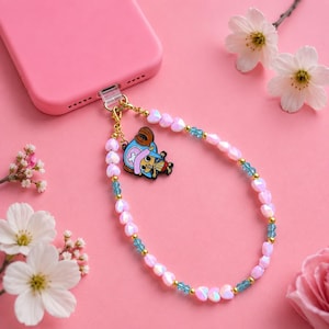 May include: A pink phone case with a beaded phone charm. The charm features a cartoon character pendant and a beaded chain of pink heart-shaped beads, gold beads, and blue beads. The background is a solid pink colour with white flowers.